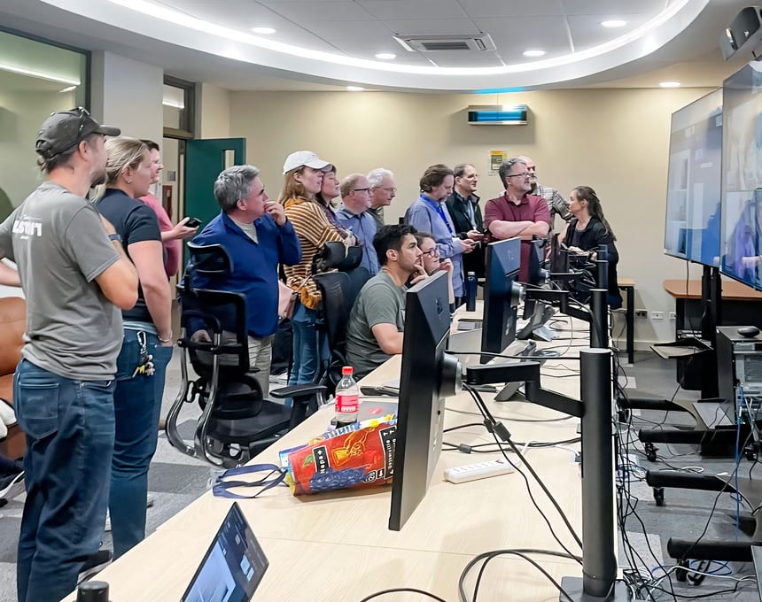 A team gathers around wall-mounted and desktop screens in a bright operations room, reflecting collaborative operational decision-making.